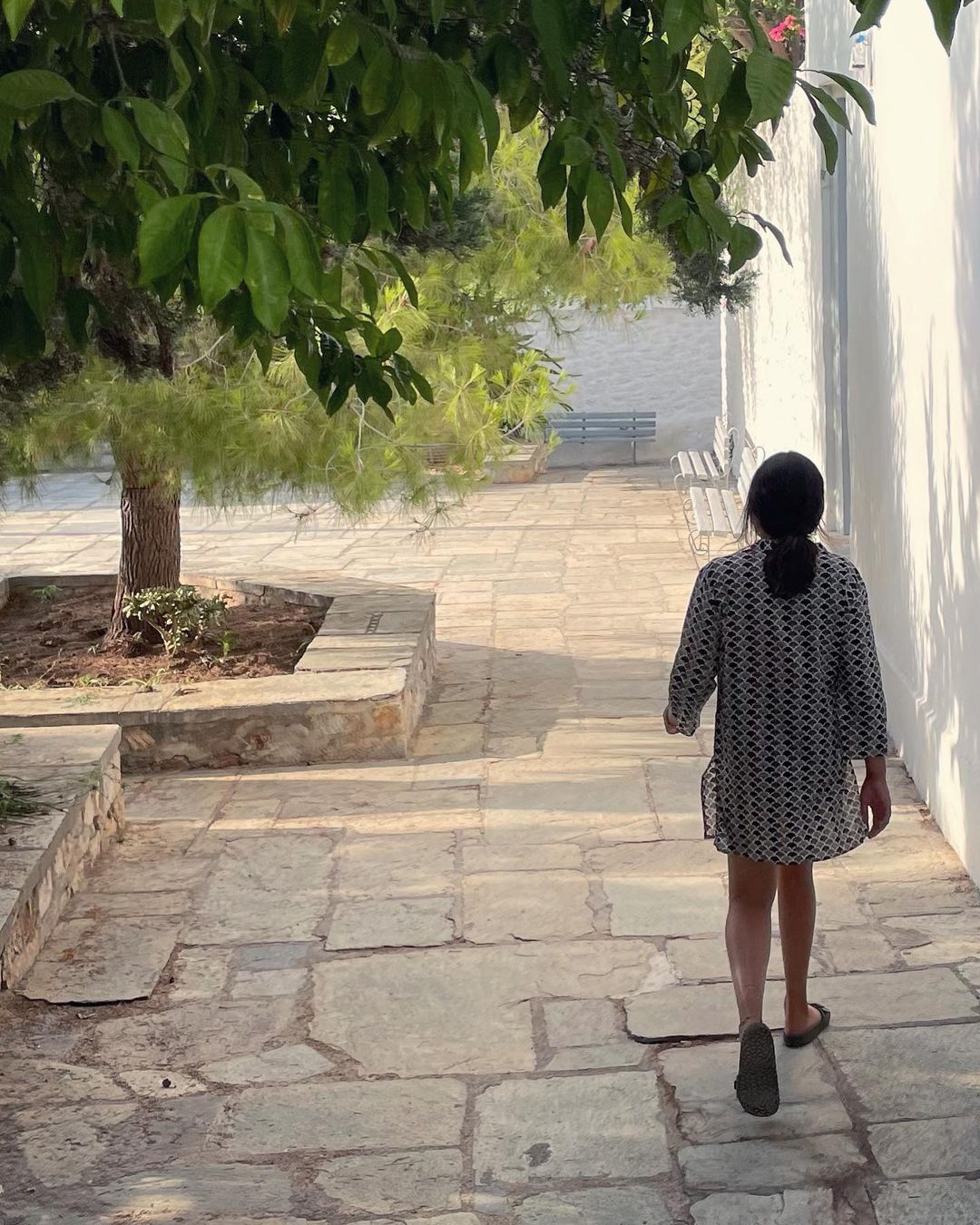 Back view of a child walking on a stone path next to a white wall, with a tree providing shade and sunlight creating shadows.