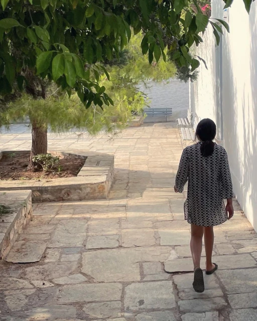 Back view of a child walking on a stone path next to a white wall, with a tree providing shade and sunlight creating shadows.