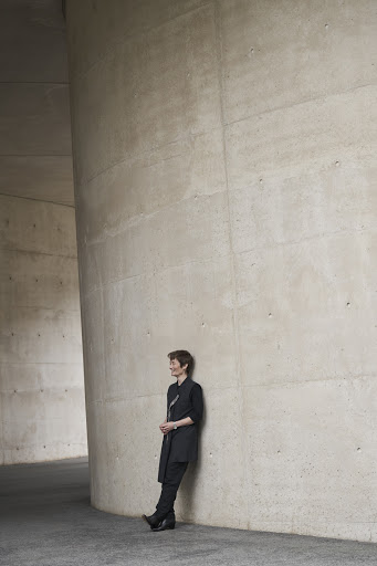 Adult woman in a black outfit leans against a modern grey concrete column inside an architectural structure with neutral, soft lighting.