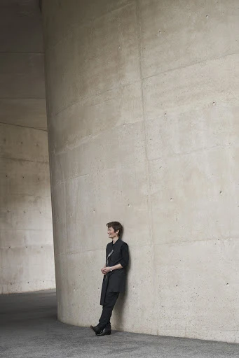 Adult woman in a black outfit leans against a modern grey concrete column inside an architectural structure with neutral, soft lighting.