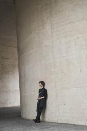 Adult woman in a black outfit leans against a modern grey concrete column inside an architectural structure with neutral, soft lighting.