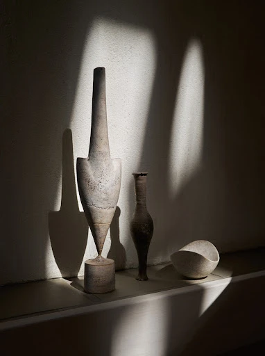 Still life photograph featuring a collection of sculptural vases and vessels in neutral tones on a shelf, bathed in light and shadow.