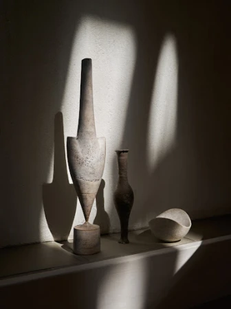 Still life photograph featuring a collection of sculptural vases and vessels in neutral tones on a shelf, bathed in light and shadow.