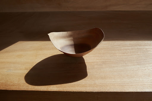 Close-up of a minimalist, light-brown wooden bowl, partially lit by sunlight, sitting on a matching wooden surface.