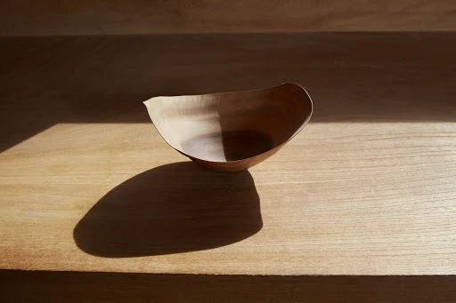 Close-up of a minimalist, light-brown wooden bowl, partially lit by sunlight, sitting on a matching wooden surface.