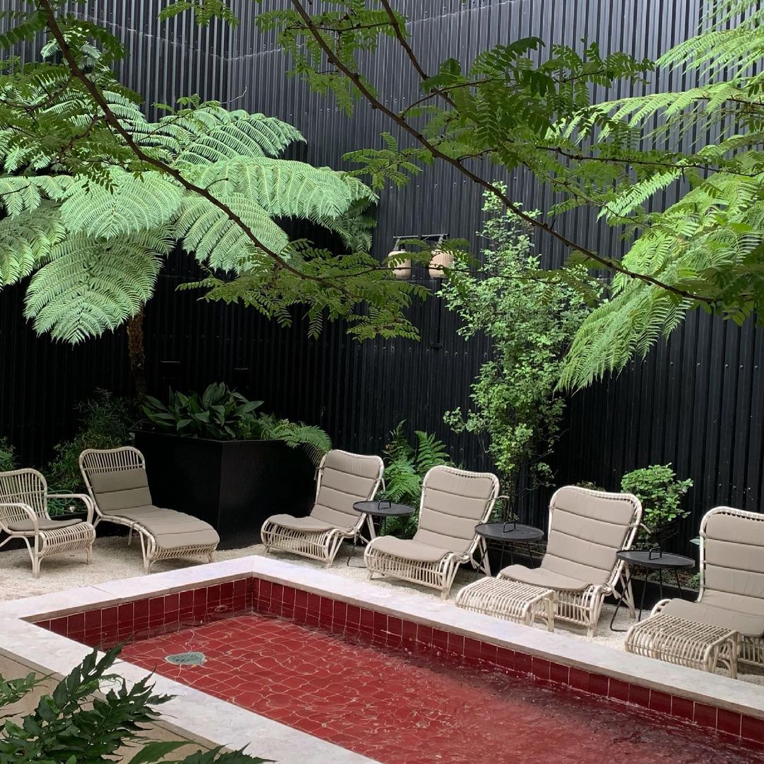 Outdoor relaxation area featuring wicker lounge chairs arranged around a small, tiled pool, surrounded by lush greenery and trees.