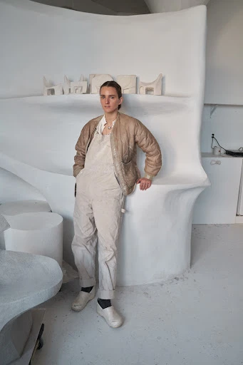 Woman in beige jacket and overalls poses in an all-white artist's studio with organic modern design elements and art objects.