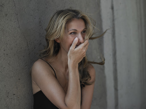 Candid portrait of a blonde woman laughing with her hand covering her mouth against a textured concrete wall.