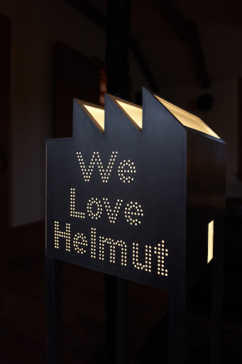 Illuminated sign in the shape of a factory with the text 'We Love Helmut' against a dark background.