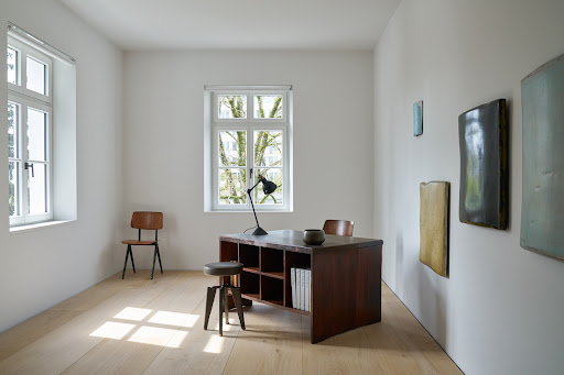 Minimalist interior with a dark wooden desk, black lamp, small stool, and abstract artworks hanging on a white wall.