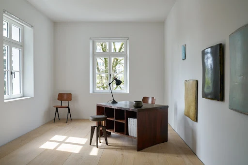 Minimalist interior with a dark wooden desk, black lamp, small stool, and abstract artworks hanging on a white wall.