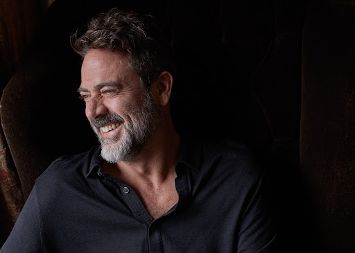 Portrait of actor Jeffrey Dean Morgan smiling while seated in a dark armchair, wearing a dark buttoned shirt.