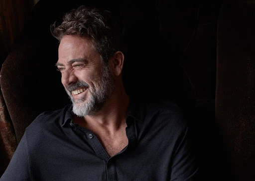 Portrait of actor Jeffrey Dean Morgan smiling while seated in a dark armchair, wearing a dark buttoned shirt.