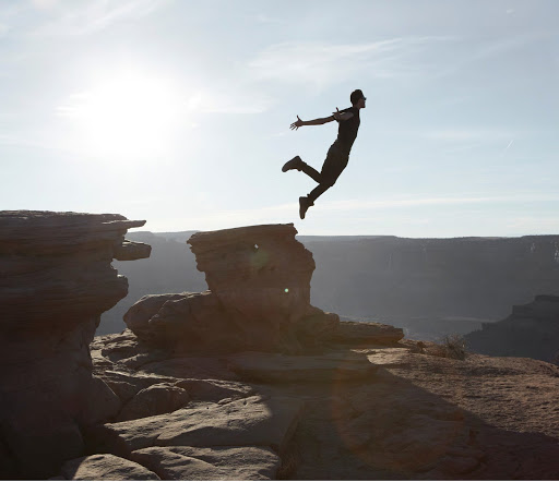 Silhouette of a man leaping between rocky cliff ledges against a pale sky captured by Martien Mulder