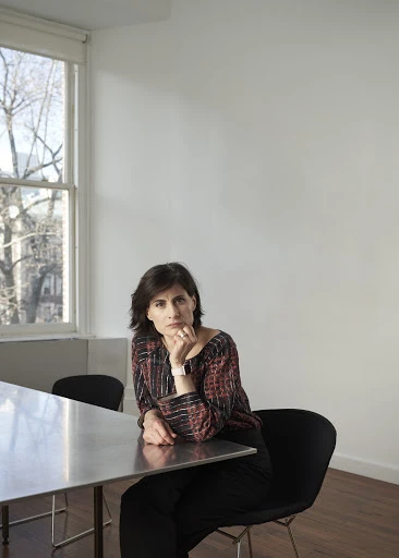 Indoor portrait of a woman with dark hair sitting at a table in front of a window wearing a patterned top and dark pants.