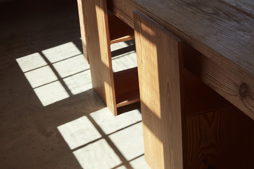 Angled shot of a wooden table and shelf with window-shaped shadows cast on the floor, creating a warm and inviting interior scene.