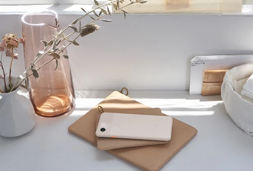 Minimalist product photography of a smartphone, vase, and floral arrangement on a white surface in front of a window.