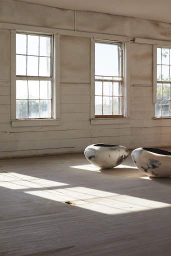 Two ceramic vessels on a wooden floor in a sunlit room with white walls and windows create a serene and minimal art installation.