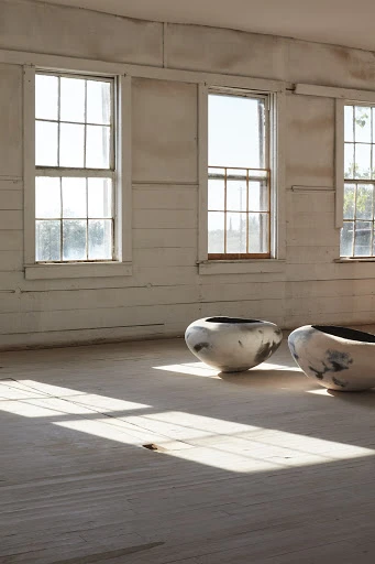 Two ceramic vessels on a wooden floor in a sunlit room with white walls and windows create a serene and minimal art installation.