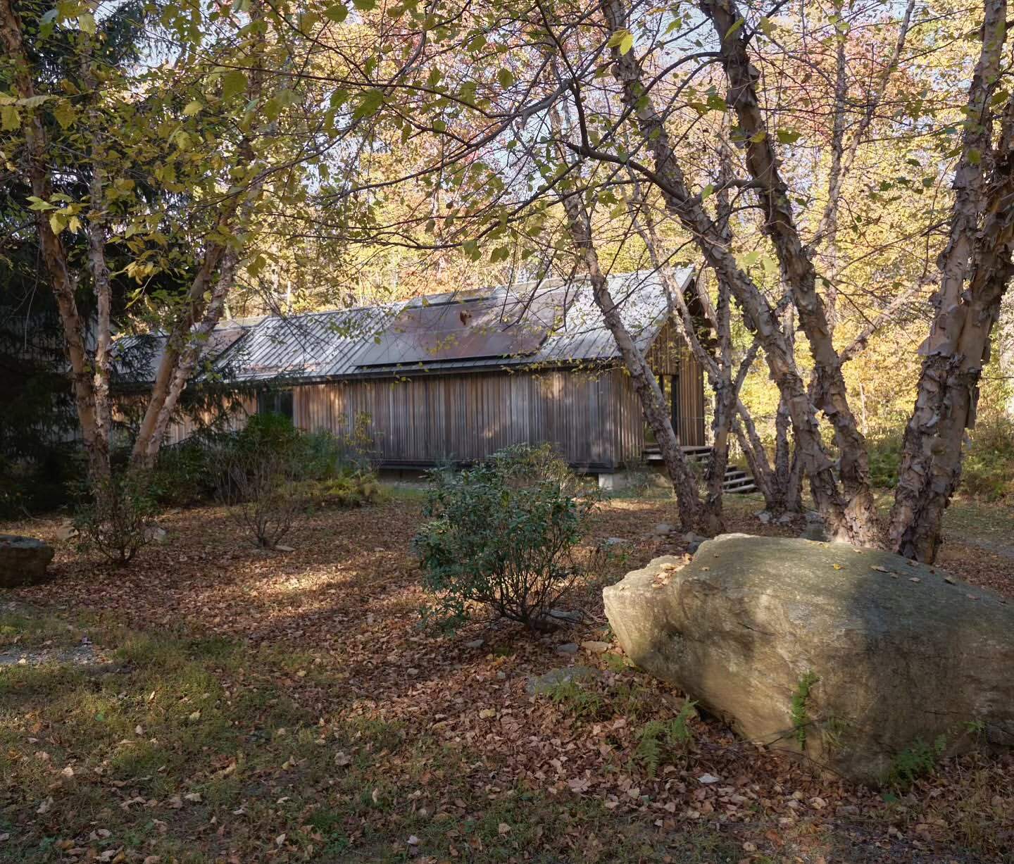 Exterior view of a weathered wood building surrounded by trees with autumn foliage in a rural environment on a sunny day.