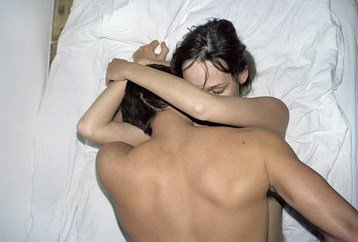 Top-down view of a man and a woman embracing in bed under white sheets in a softly lit indoor environment, capturing a moment of intimacy.