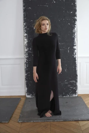 Woman in long black dress standing against a dark weathered panel inside a room with herringbone wood flooring