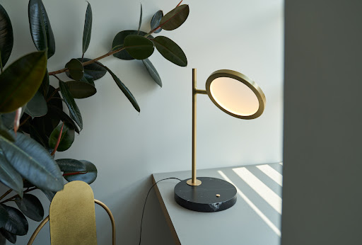 Interior with a stylish gold desk lamp featuring a circular LED and a black marble base next to a ficus plant.
