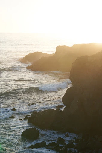 Ocean waves crash against rocky cliffs at sunset with sunflare, creating a scenic coastal landscape with golden light and natural beauty.