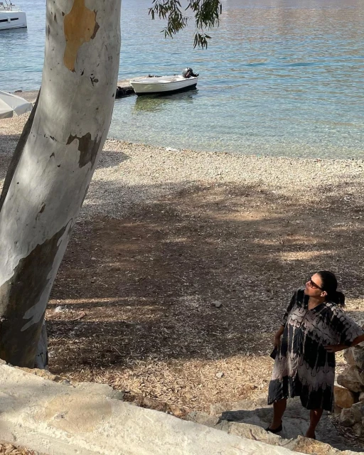 Woman in a dress stands on stone steps near a beach with clear water and a small boat in the background, near a large tree trunk.