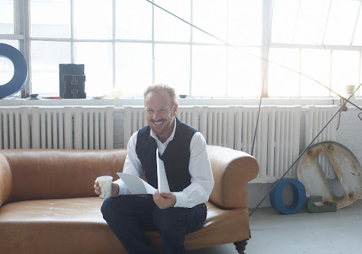 Smiling man in vest sits on leather couch in bright room, holding papers and a drink, with large letter art in background.
