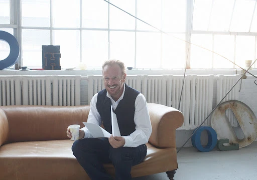 Smiling man in vest sits on leather couch in bright room, holding papers and a drink, with large letter art in background.