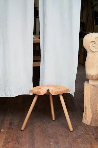 Handmade wooden stool with three legs, displayed in a workshop setting with wooden head sculpture and white fabric backdrop.