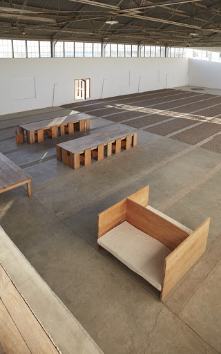 Overhead shot of a large empty industrial space featuring geometric wooden tables and a small wooden sleeping structure.