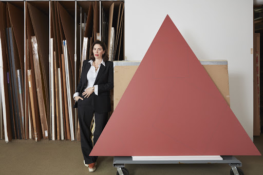 A woman in a suit stands next to a large red triangle leaning on a dolly inside what appears to be a workshop or art studio.