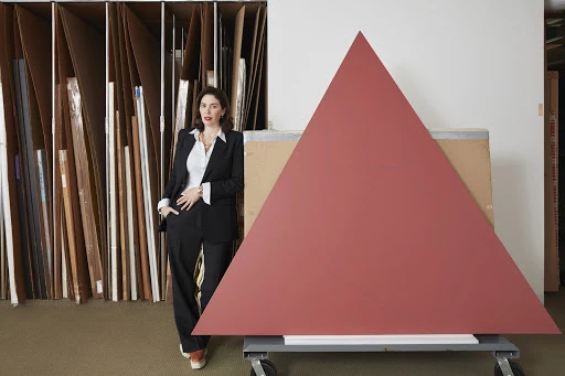 A woman in a suit stands next to a large red triangle leaning on a dolly inside what appears to be a workshop or art studio.