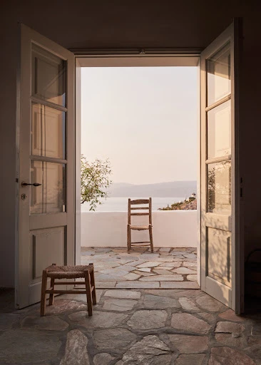 View of a stone patio and coastal landscape through open white double doors, with a wooden chair and stool on the patio.
