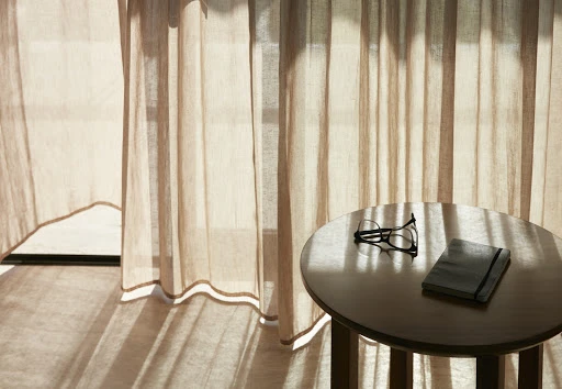 Still life of eyeglasses and notebook on a round table against neutral beige curtains in sunlight, creating a soft and tranquil scene.