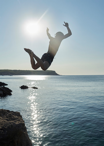 A person jumps from a cliff into the sea on a sunny day. The figure is silhouetted against the bright sun, with rocky cliffs visible.