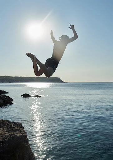 A person jumps from a cliff into the sea on a sunny day. The figure is silhouetted against the bright sun, with rocky cliffs visible.