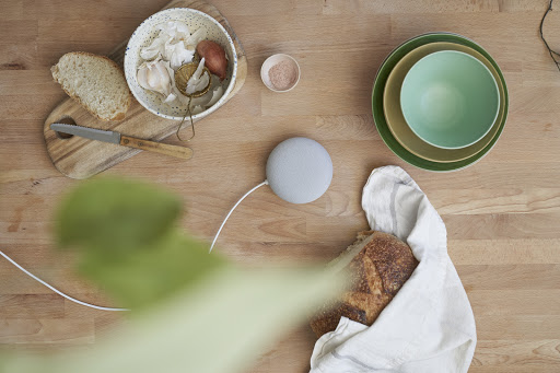 Overhead shot of artisan bread, bowl of garlic, shallots, digital assistant device, and green bowls on a wooden kitchen table.