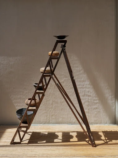 Rustic metal ladder shelves with vintage pottery arranged in front of neutral wall in a minimalist interior with wooden floors.