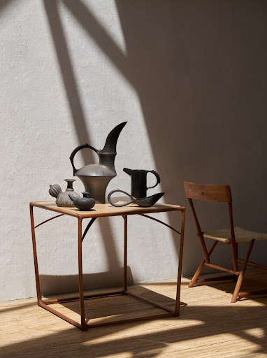 Still life featuring a collection of ceramic vessels displayed on a small table, illuminated by natural light casting shadows on a wall.