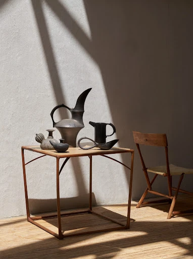 Still life featuring a collection of ceramic vessels displayed on a small table, illuminated by natural light casting shadows on a wall.
