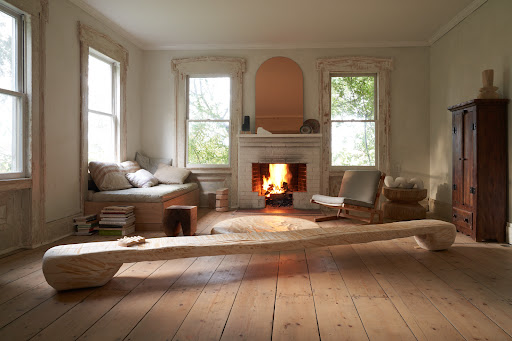 Living room with wooden floors, fireplace, and windows providing natural light, complemented by a wooden bench and minimalist decor.