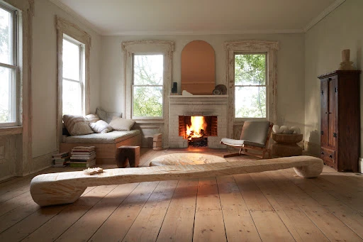 Living room with wooden floors, fireplace, and windows providing natural light, complemented by a wooden bench and minimalist decor.