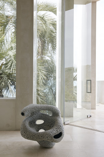 Interior view with sculptural porous chair on tiled floor, flanked by a window showing palm trees, next to a glass-walled hallway.