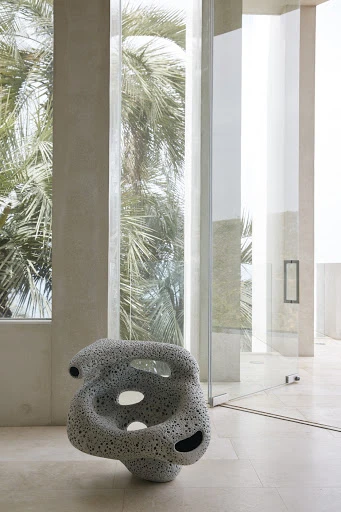 Interior view with sculptural porous chair on tiled floor, flanked by a window showing palm trees, next to a glass-walled hallway.