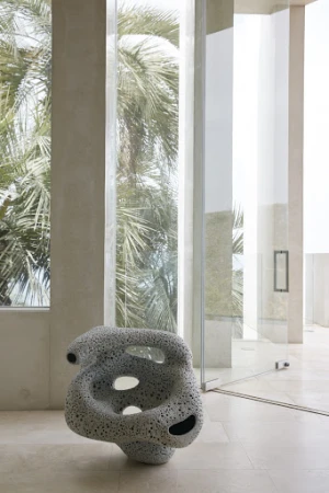 Interior view with sculptural porous chair on tiled floor, flanked by a window showing palm trees, next to a glass-walled hallway.