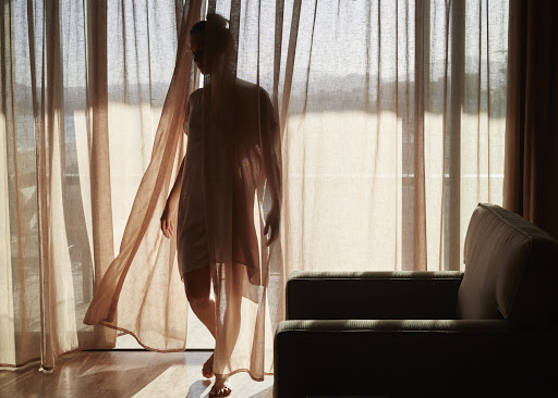 Silhouette of a slender woman in a nightgown standing near a curtain-covered window, with an armchair visible in a dimly lit room.