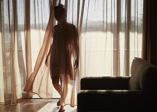 Silhouette of a slender woman in a nightgown standing near a curtain-covered window, with an armchair visible in a dimly lit room.